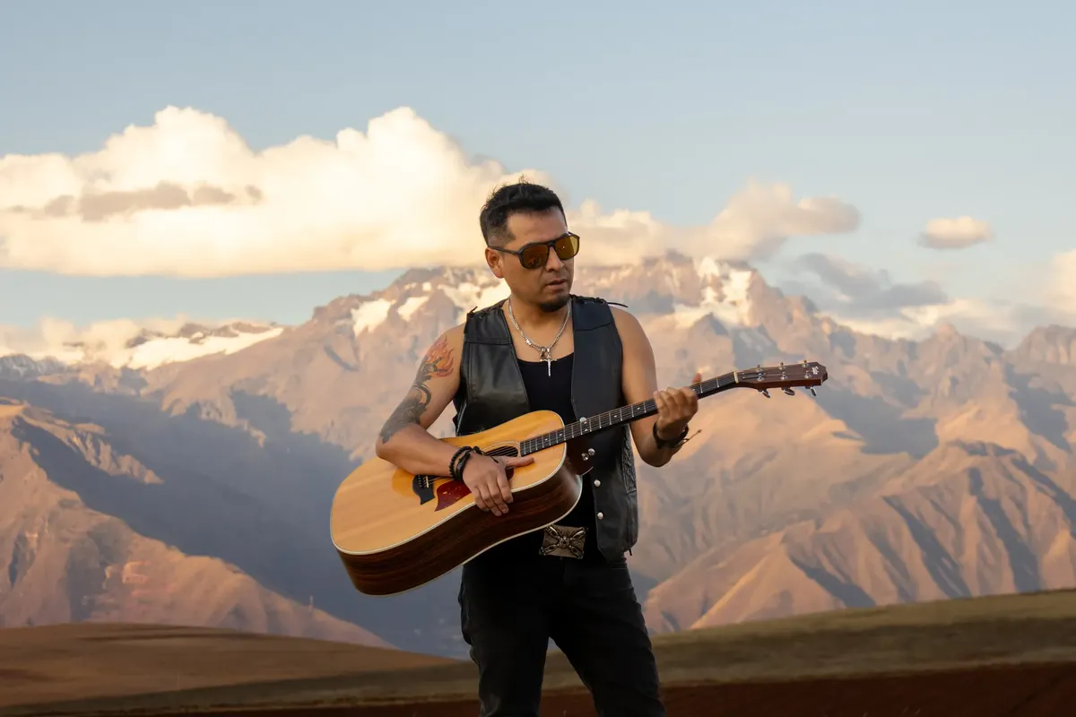 Guitarist wearing sunglasses plays acoustic guitar in front of majestic mountain backdrop during daylight.